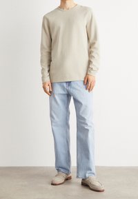 Beige långärmad skjorta, ljusblå raka jeans och grå loafers med röd sula. Minimalistisk design och subtila texturdetailler.