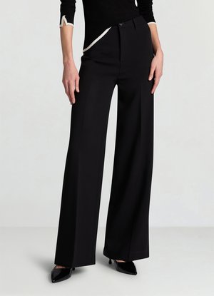 Pantalon large noir en tissu léger. Caractérisé par une taille haute, des plis devant et une coupe ajustée, porté avec des talons noirs pointus.