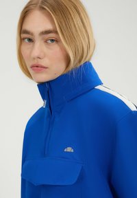 Blauwe hooggecollar jacket met een ritssluiting in de nek, voorzak en witte accentstrepen op de schouders. Glad textuur en casual ontwerp.