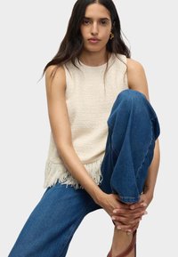 Top di colore crema con una finitura frangiata, abbinato a jeans in denim blu. Modella seduta, con un ginocchio sollevato, indossa sandali marroni con cinturini.