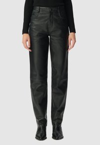 Vrouw in hoge zwarte leren broek met voorzakken, gecombineerd met een zwart geribbelde lange mouwen top en zwarte pointed schoenen.