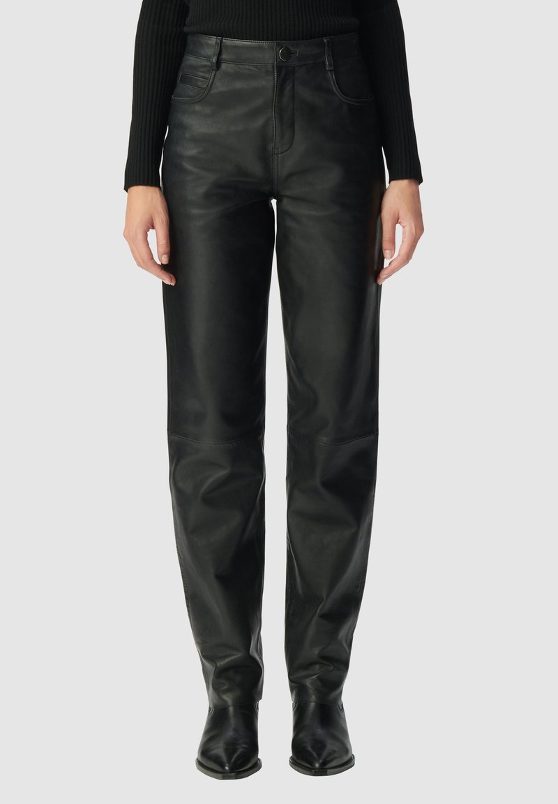 Vrouw in hoge zwarte leren broek met voorzakken, gecombineerd met een zwart geribbelde lange mouwen top en zwarte pointed schoenen.