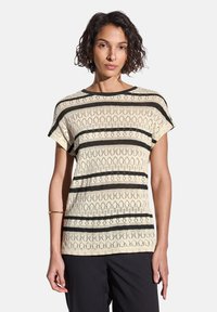 Beige short-sleeve top with intricate black striped patterns and open-knit texture. Round neckline, relaxed fit, paired with black trousers.