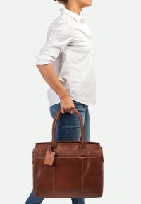 Braune Lederhandtasche mit strukturiertem Design, doppelten Griffen und einem Gepäckanhänger. Verfügt über eine glatte Textur und minimalistische Akzente.