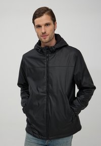 Giacca nera impermeabile con cappuccio, dotata di zip, tasche laterali e polsini elasticizzati, realizzata in materiale liscio e lucido.