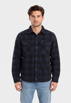 Giovane uomo con capelli corti castani che indossa una camicia a quadri nera e blu abbottonata e jeans azzurri, in piedi contro uno sfondo semplice.