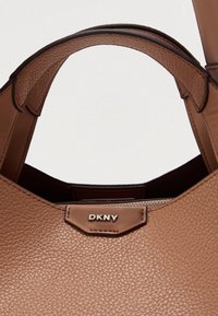 Barna szemcsés bőrből készült kézitáska sima bőrből készült szegéllyel, cipzáras záródással és az elülső oldalon jól látható DKNY logóval.