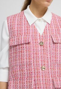 Růžový tvídový vest, s texturovaným povrchem, který má dvě náprsní kapsy a stříbrný knoflík. Nesu na bílou košili s límečkem.