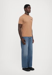 Brun kortermet t-skjorte med en liten logo på brystet, kombinert med løse blå denimjeans og sorte joggesko.