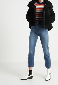 Sort fuzzy jakke med lynlås, flerfarvet stribet sweater, lyseblå lige jeans, og hvide ankelstøvler med sorte accenter.