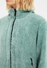 Giacca di pile azzurro chiaro con una superficie testurizzata e soffice, colletto alto e dettagli con zip nera. Presenta una tasca laterale con chiusura a zip.