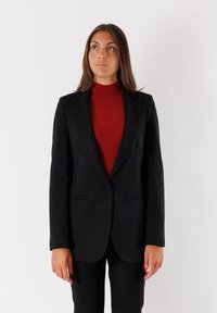 Blazer nero con un solo bottone, rever a rotondo e due tasche frontali, indossato su un dolcevita rosso a coste, con vestibilità sartoriale.