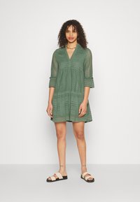 Green lace dress with a V-neckline, three-quarter sleeves, and a flared skirt featuring patterned detailing. Paired with beige sandals.