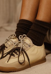 Πόδια με σκουρόκαφες ριγωτές κάλτσες και μπεζ αθλητικά παπούτσια με καφέ και γκρι κορδόνια, που ακουμπούν σε ένα φωτεινό χαλι.