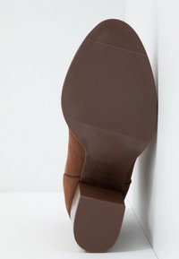 Brązowy zamszowy botek za kostkę na słupku z warstwowaną podeszwą z gumy o fakturze i subtelnym przeszyciem wzdłuż szwu.