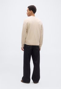 Άνδρας με πλάτη φοράει μπεζ πουλόβερ, φαρδιά μαύρα τζιν και καφέ παπούτσια μπροστά σε απλό ανοιχτό φόντο.