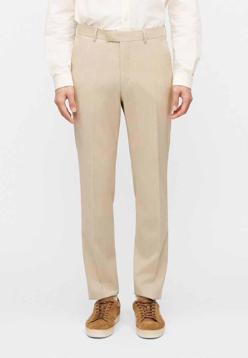 Μπεζ παντελόνι σε ίσια γραμμή από ελαφρύ ύφασμα, με επίπεδη μπροστινή όψη και θηλιές για ζώνη. Συνδυάζεται με καφέ σουέντ αθλητικά παπούτσια.