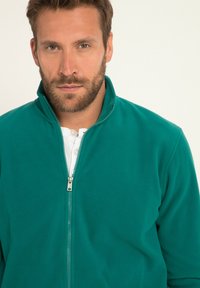 Giacca in pile color teal con colletto alto e zip completa, caratterizzata da una texture liscia e una camicia bianca sotto.