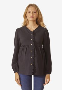 Frau mit langen blonden Haaren, die eine schwarze Bluse mit langen Ärmeln und Rüschensaum sowie goldenen Knöpfen trägt, kombiniert mit blauen Jeans, steht aufrecht.