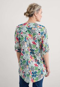 Koszulka w kwiatowy wzór z wyrazistymi różowymi, zielonymi i niebieskimi motywami na jasnym tle; ma okrągły dekolt i rękawy o długości trzech ćwiartek.