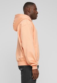 Sudadera de melocotón hecha de un tejido suave, con una capucha grande y puños de canalé. Combinada con jeans gris oscuro, que muestran un ajuste relajado.