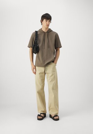 Sudadera con capucha marrón, pantalones beige claros y sandalias negras. La sudadera tiene mangas cortas y un bolsillo delantero; los pantalones son de corte holgado.