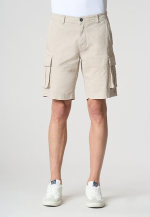 Pantaloni cargo beige in cotone, con tasche laterali, parte anteriore piatta e chiusura con bottone. Indossati con sneakers bianche.