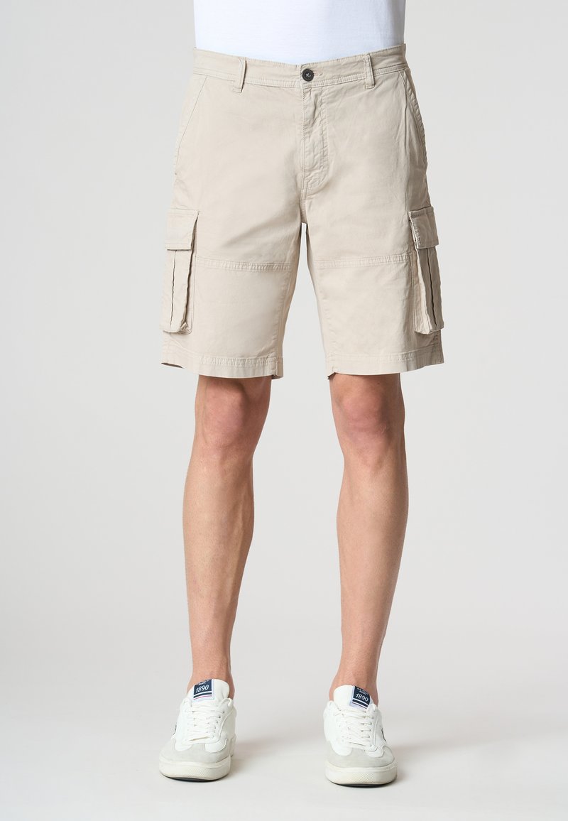 Pantaloni cargo beige in cotone, con tasche laterali, parte anteriore piatta e chiusura con bottone. Indossati con sneakers bianche.