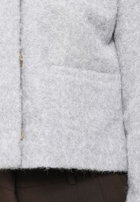 Gros plan d'une veste en laine gris clair texturée avec poche plaquée devant et boutons, portée sur un pantalon marron foncé.