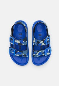 Siniset sandaalit, joissa on camo-nauhat ja kaksi säädettävää solkea. Texturoitu pohja tarjoaa paremman pidon; pehmeä materiaali mukavuutta varten.