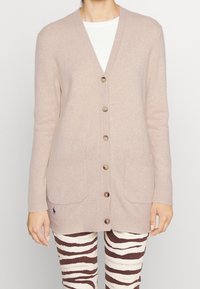 Kvinna som bär en beige knappad cardigan med framfickor över en vit topp och bruna och vita randiga byxor.