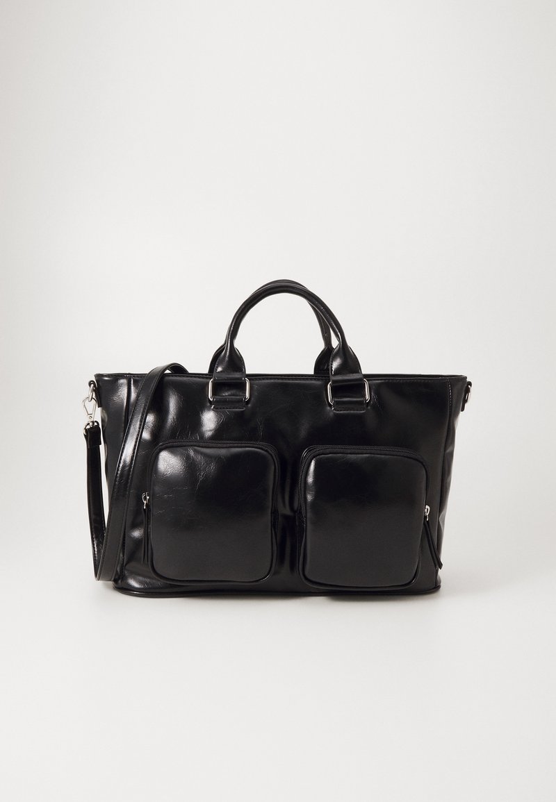 Sac cabas en cuir noir avec deux poches avant zippées, double poignée supérieure et une bandoulière détachable, sur un fond clair uni.