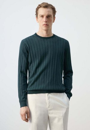 Hombre con jersey de punto oscuro verde de manga larga y pantalones blancos, de pie contra un fondo claro y liso, con las manos relajadas.