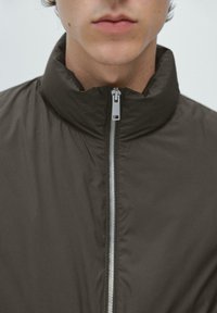 Ciemnobrązowa kurtka z wysokim kołnierzem i srebrnymi detalami zamka błyskawicznego. Materiał wydaje się gładki i lekko pikowany.