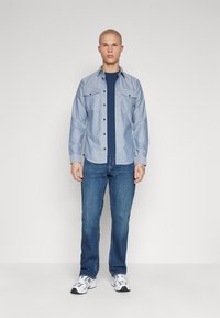 Chemise bleu clair à manches longues avec deux poches poitrine, portée sur un t-shirt marine, associée à un jean bleu et des baskets blanches. Fond uni.