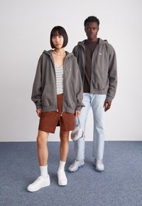 Grå zip-up hættetrøje, stribet bluse, brun nederdel; model i hvide sneakers og iført en halskæde. Anden model i en brun skjorte og lyse jeans, grå sko.