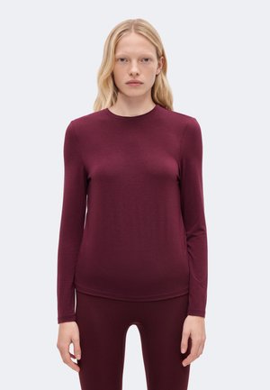 Un mannequin blond se tient devant un fond clair, vêtu d’un haut à manches longues bordeaux et d’un legging assorti.