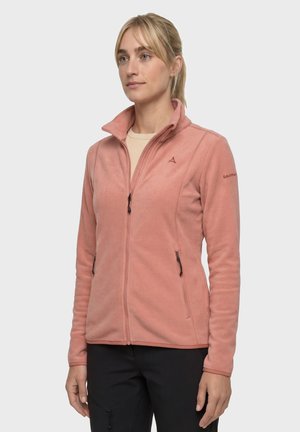 Frau trägt eine hellrosa Fleecejacke mit Reißverschluss und hohem Kragen sowie schwarze Hose, steht vor einem schlichten hellen Hintergrund.