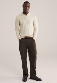 Ein Mann steht mit in die Taschen gesteckten Händen und trägt ein beiges Langarm-Poloshirt, dunkelbraune Hosen, einen braunen Gürtel und dunkelbraune Loafers vor neutralem Hintergrund.