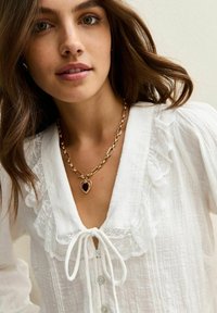 Jonge vrouw met bruin haar draagt een witte blouse met knopen en kantafwerking, en een gouden ketting met een donker hartvormig hanger.