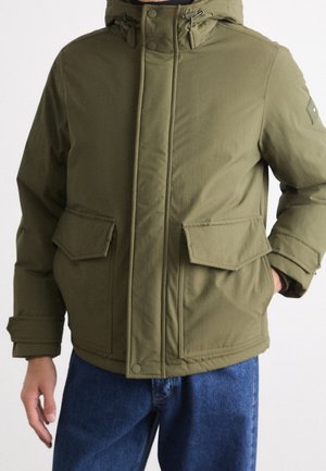 Mann trägt olivgrüne Kapuzen-Winterjacke mit vorderen Klappentaschen und blaue Jeans, Hand in der Tasche.