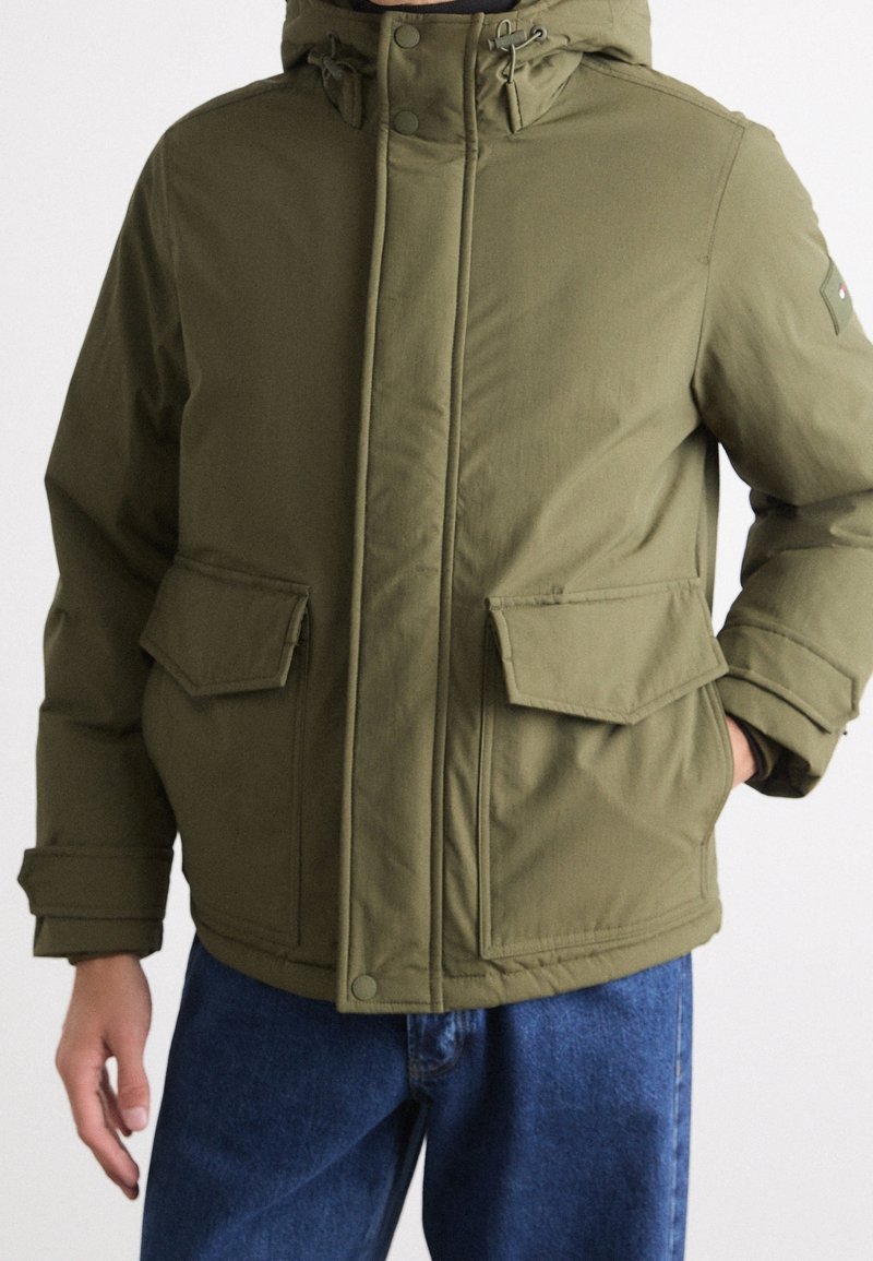 Homme portant une veste d'hiver à capuche vert olive avec des poches avant à rabat et un jean bleu, main dans la poche.