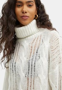 Maglione bianco a cretti con colletto alto a coste. La trama aperta rivela lo strato sottostante. Texture morbida e silhouette rilassata.