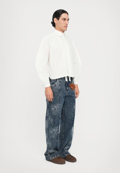 Camisa blanca de cuello con un diseño recortado, combinada con pantalones vaqueros denim azul sueltos con bordados florales. Zapatos marrones completan el atuendo.