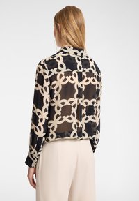 Blusa nera trasparente con un motivo a catena astratto beige, maniche lunghe e dettaglio sul retro. Il tessuto sembra leggero e fluido.