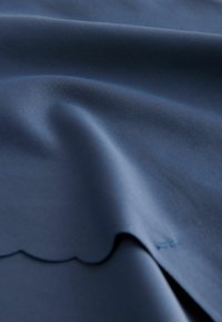 Material moale albastru cu textură fină, pliuri delicate și margine cu detaliu ondulat, prezentat în prim-plan.