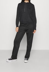 Černá mikina s kapucí a texturovaným povrchem, kombinovaná s černými hladkými kalhotami a klasickými černými teniskami s bílými podrážkami a tkaničkami.