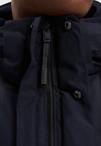 Tamsiai mėlynas pūkinis švarkas su aukštu apykakle, turintis priekinį užtrauktuką, užsegamas mygtukais ir tekstūruota medžiaga. Ant užtrauktuko matoma prekės ženklo etiketė.