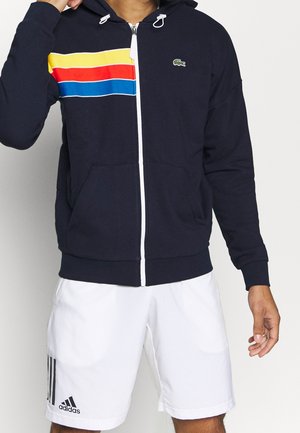 Mies, joka käyttää tummansinistä vetoketjullista hupparia, jossa on keltaisia, punaisia ja sinisiä raitoja, sekä valkoisia Adidas-shortseja mustalla logolla ja raidoilla.