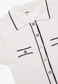 Polo shirt bianco con accenti neri, texture a coste, due tasche sul petto con bordatura e chiusura anteriore con bottoni decorativi.
